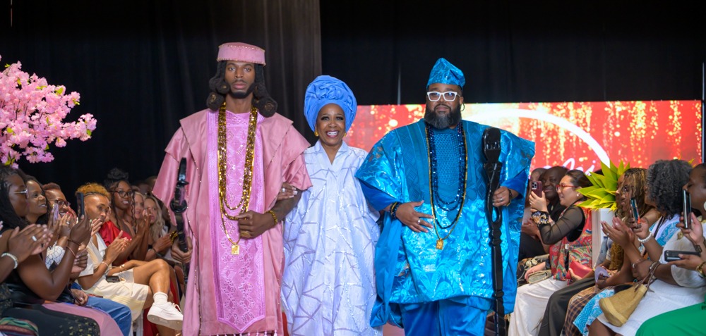 Adeyemi RoyaLabel designer Bosede Iyewarun walking the runway between two male models in pink and blue agbadas before a seated audience at Heartland International Fashion Week Season Six